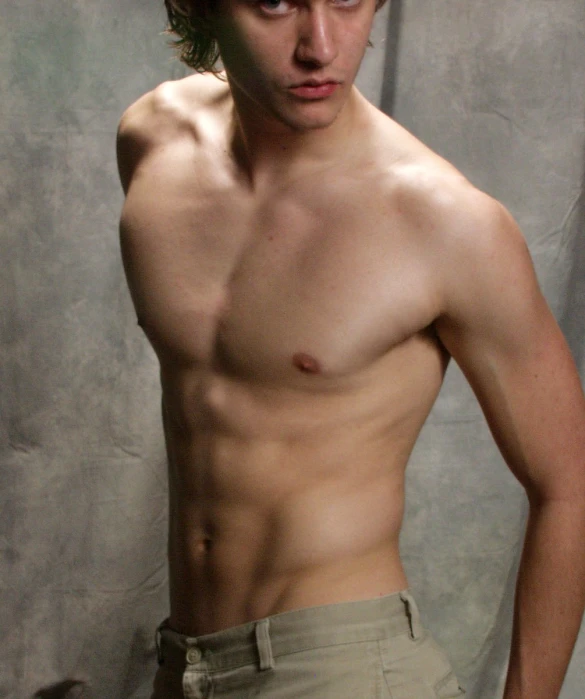 a shirtless man looks away while standing in front of a background