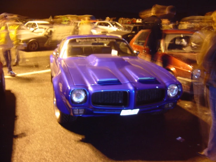 two classic muscle cars in a parking lot, with people standing around