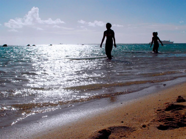 two people walking into the ocean with boats in the background