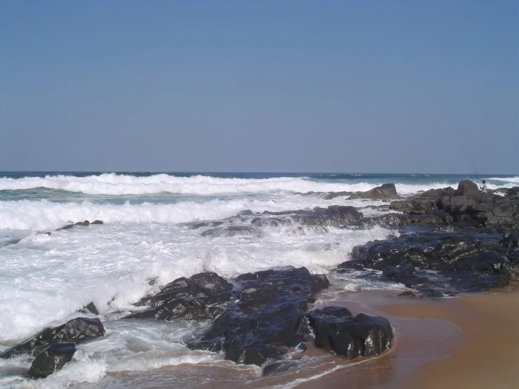 a beach with water crashing onto it, and rocks sticking out of the sand