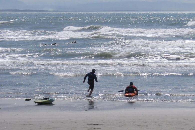 a surfer stands beside an inflatable sailboat as another walks into the water