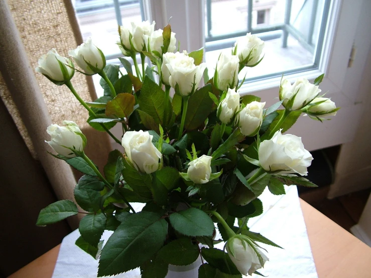 an arrangement of white roses on a table near a window