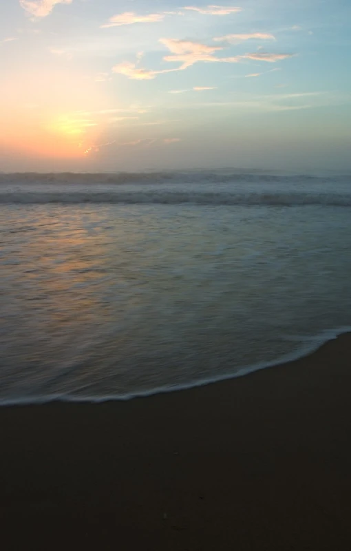 the sun sets over a sandy beach with waves and small boats in the distance