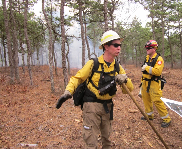 two men in yellow fire suits and hats walk on the forest path carrying wooden sticks