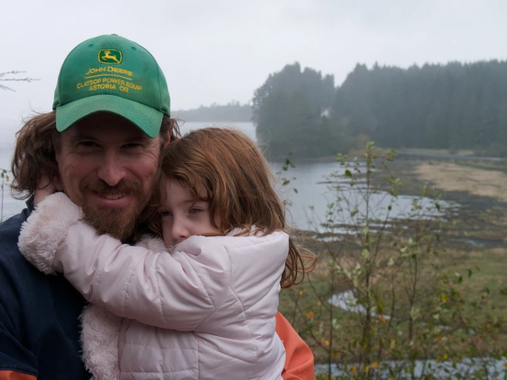 man in a green hat holding a girl with a body of water in the background