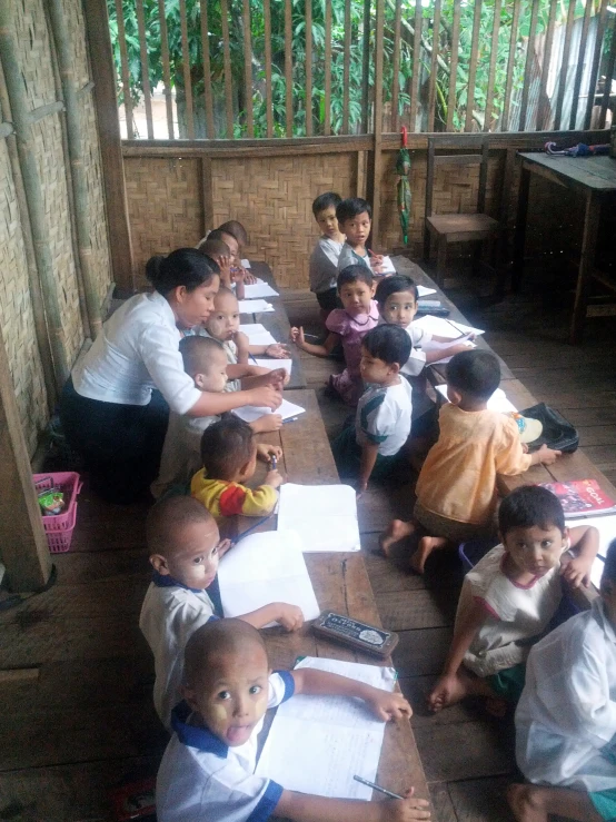 a group of children sitting at a wooden table with a woman writing