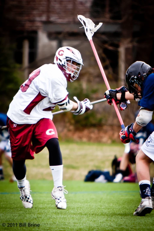 two lacrosse players wearing helmet and gloves, running towards the ball while one holds a stick