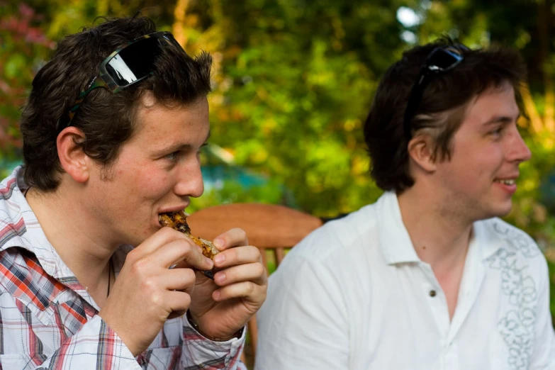 two men sit next to each other on a bench while eating