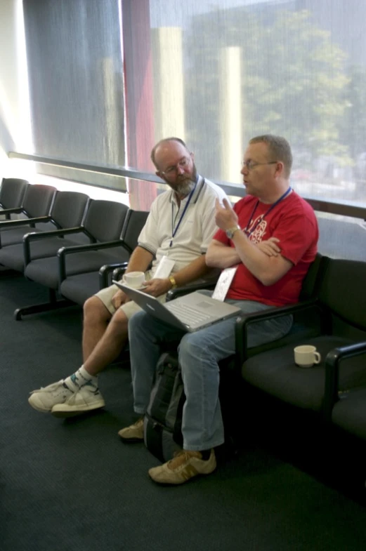 two people are sitting on an airport chair and one is holding a laptop