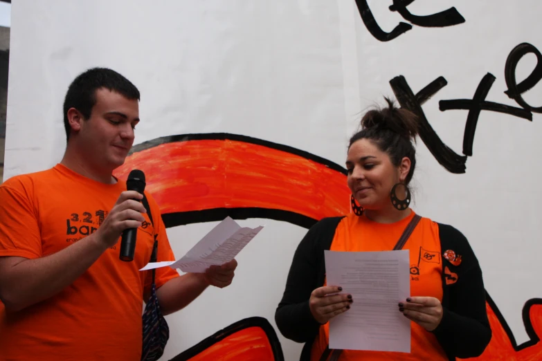 a woman with orange shirt on speaking into a microphone and two men in orange shirts