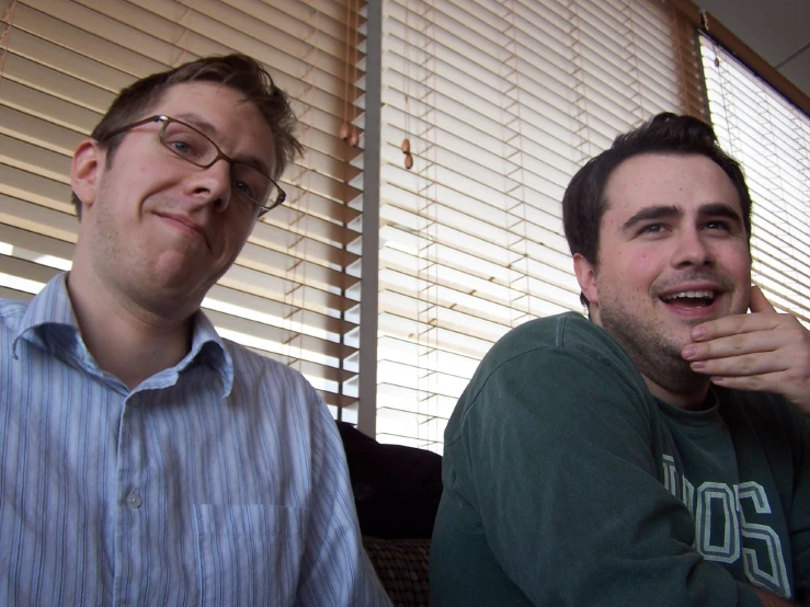 two men sitting next to each other in front of blinds