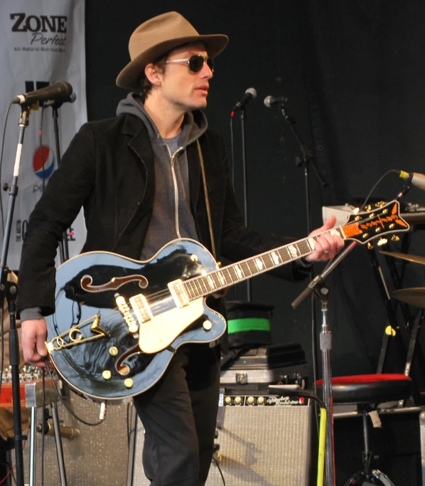 a man with sunglasses and a hat standing in front of an electric guitar