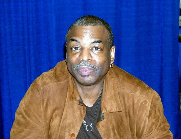 an older man sitting at a table in front of a blue curtain