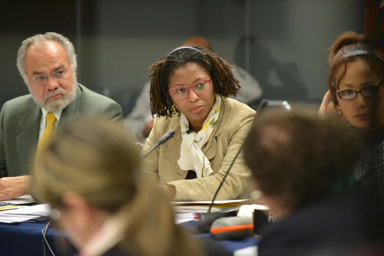 three people in a room at a table, and one is listening to the speaker