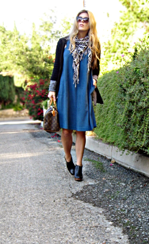 woman wearing a blue dress, black sweater and sunglasses walking down the street