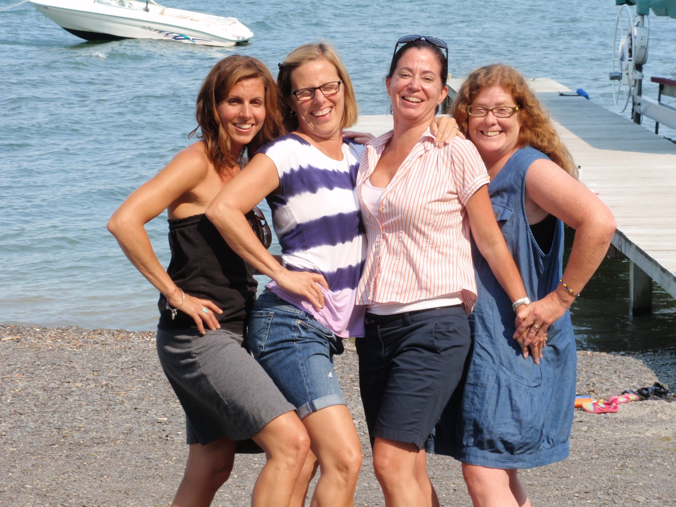 three women standing together by a lake near boats