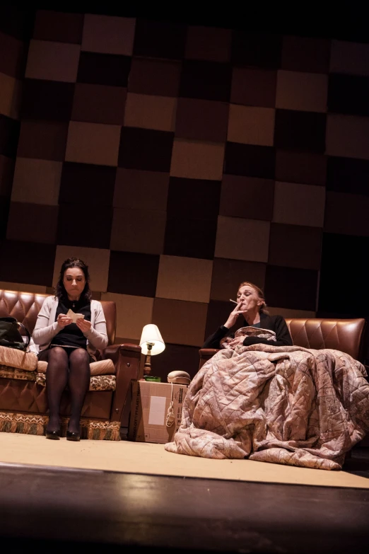 two people in chairs on stage with checkered wall behind them