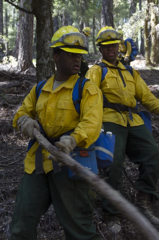 men with yellow rain suits and hats pull a rope through the woods