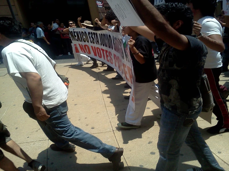 two men hold up a sign that reads no vote in front of other people walking on the street