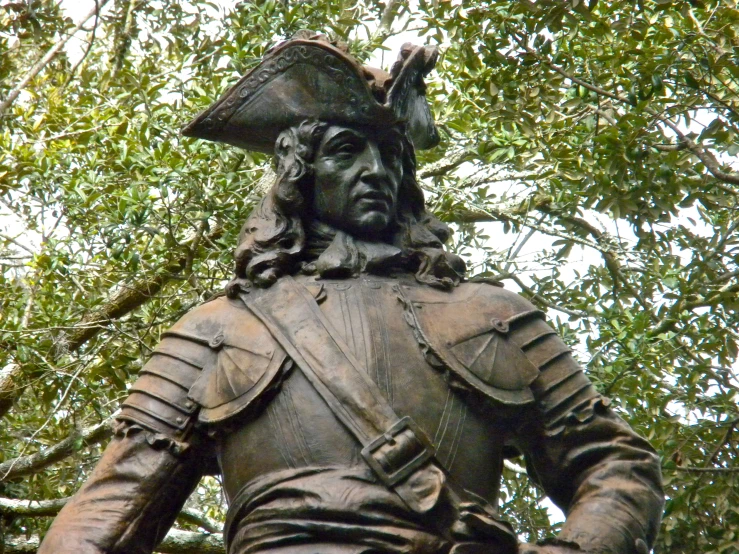 a statue of a man in armor and hat in front of trees
