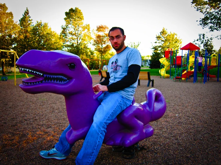 a man sitting on top of a purple toy dinosaur in a playground