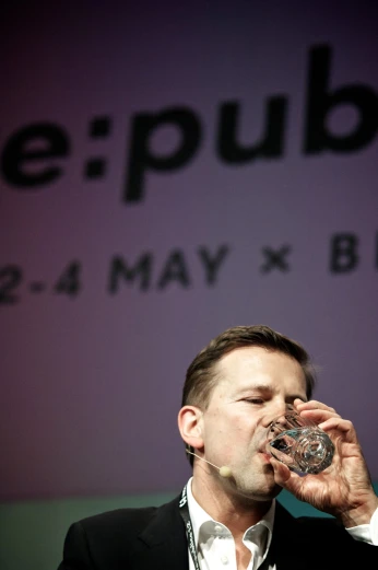 a man drinks a glass of water while talking on a stage