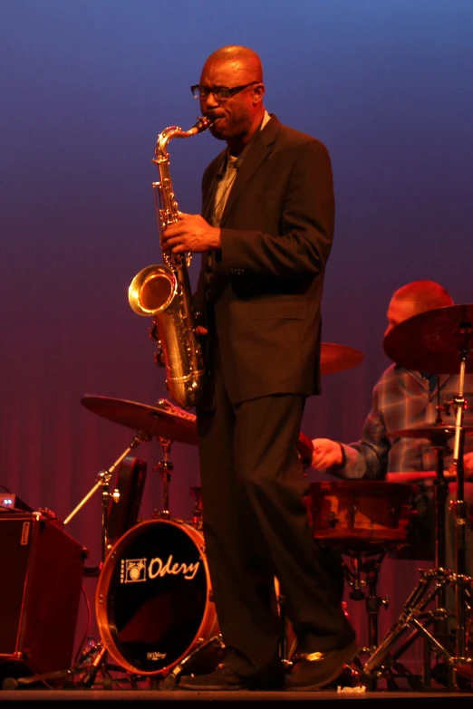 a man in a suit playing a saxophone in front of some drum kits