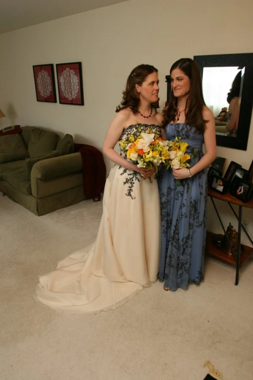 two women in formal dresses and bouquets in front of a large mirror