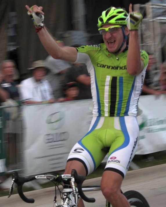 man riding bicycle with green and blue uniform waving at crowd