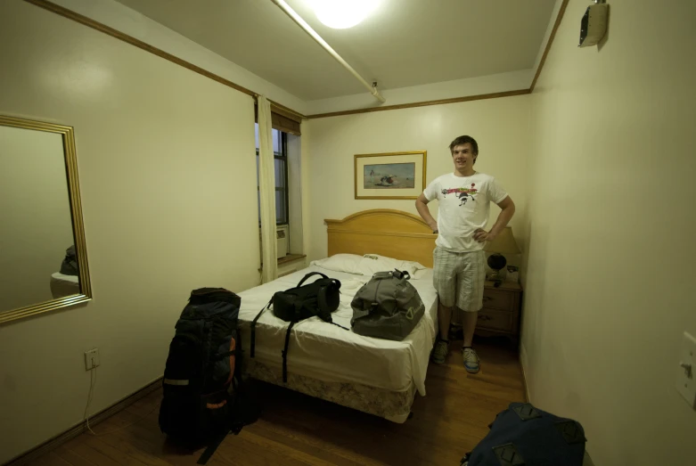 a young man standing in his room by a bed with suit cases on the bottom shelf