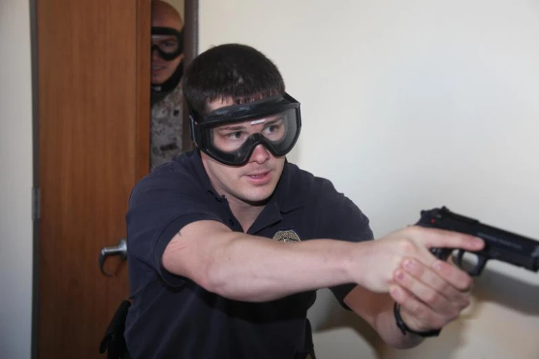 a young man dressed in black, with goggles, aiming a gun