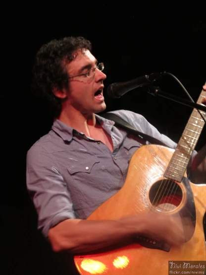 man with glasses on singing into a microphone while holding a guitar