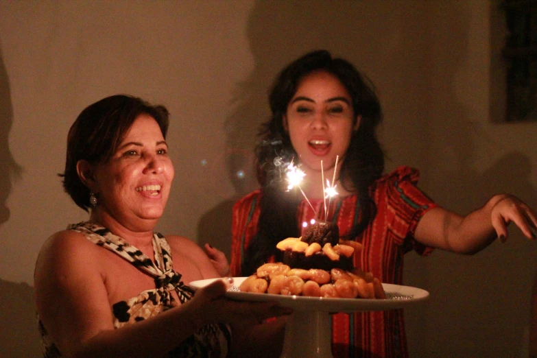 two women with lit candles on cake stand by another woman