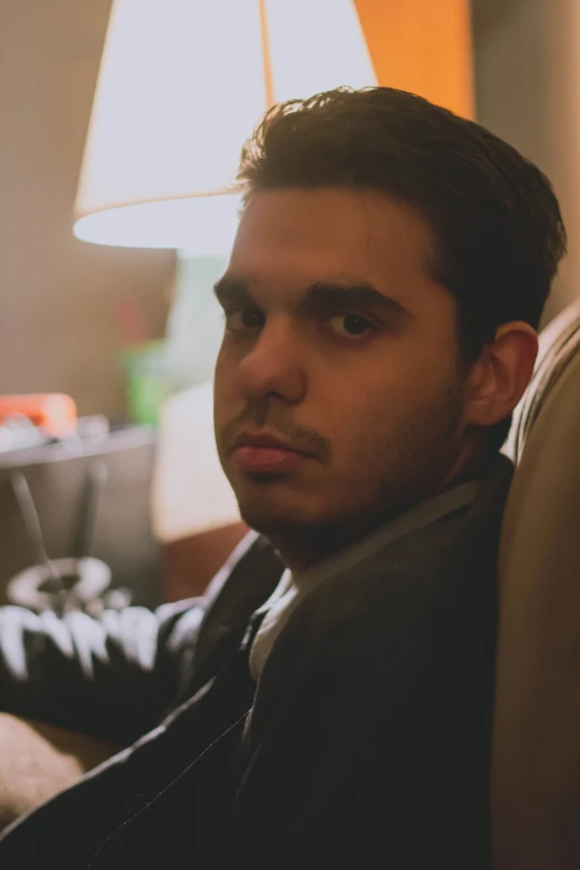 a man in a dark suit sitting next to a lamp