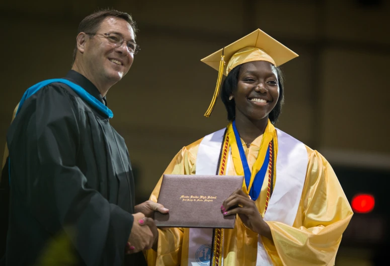 a graduate stands with his diploma in front of a smiling older graduate