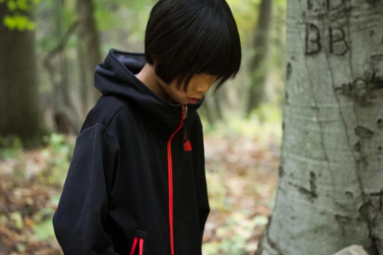 a boy wearing a black and red jacket stands in the woods