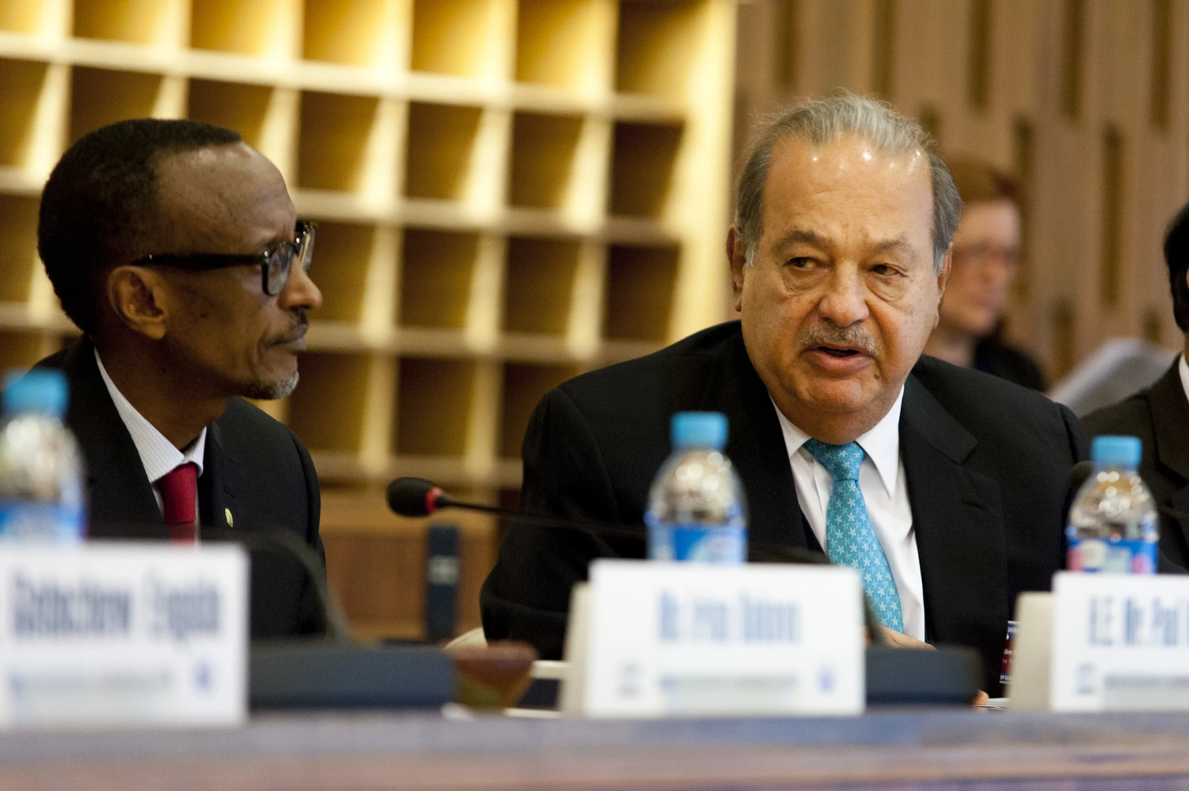 two men with business attire sit at a table with water bottles on them