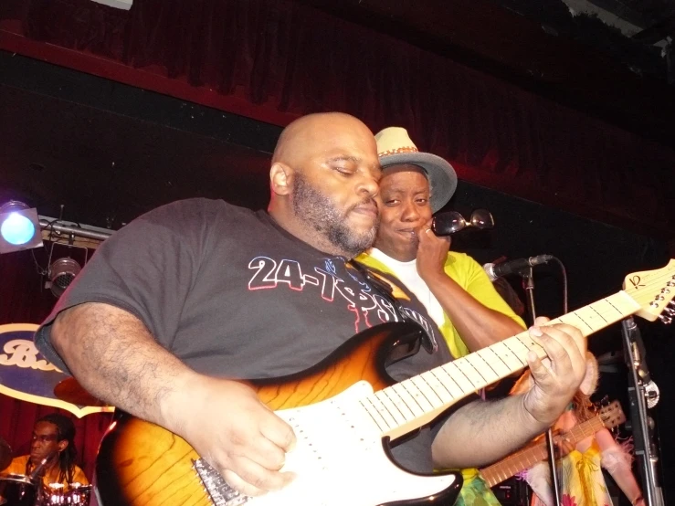 a man wearing a fedora and black t - shirt plays his guitar at a band