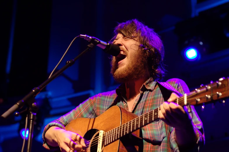 a bearded man singing and playing guitar in front of microphones