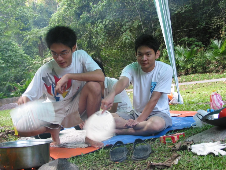 two men sit outside while making food on the ground