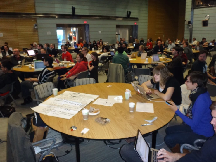 group of people gathered in a large hall with tables, chairs and laptops