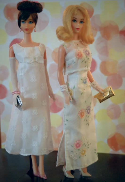 two dolls wearing white dresses and purses stand in front of a floral wall