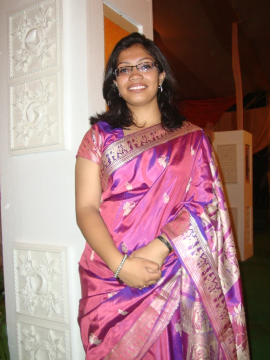 a woman standing with her arms folded wearing a pink and purple saree