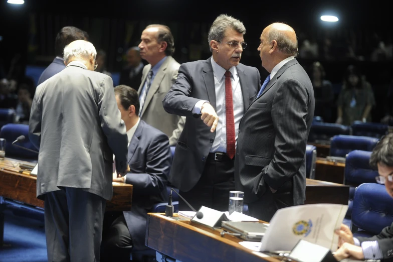 two men in suits talking to each other on the side of a table