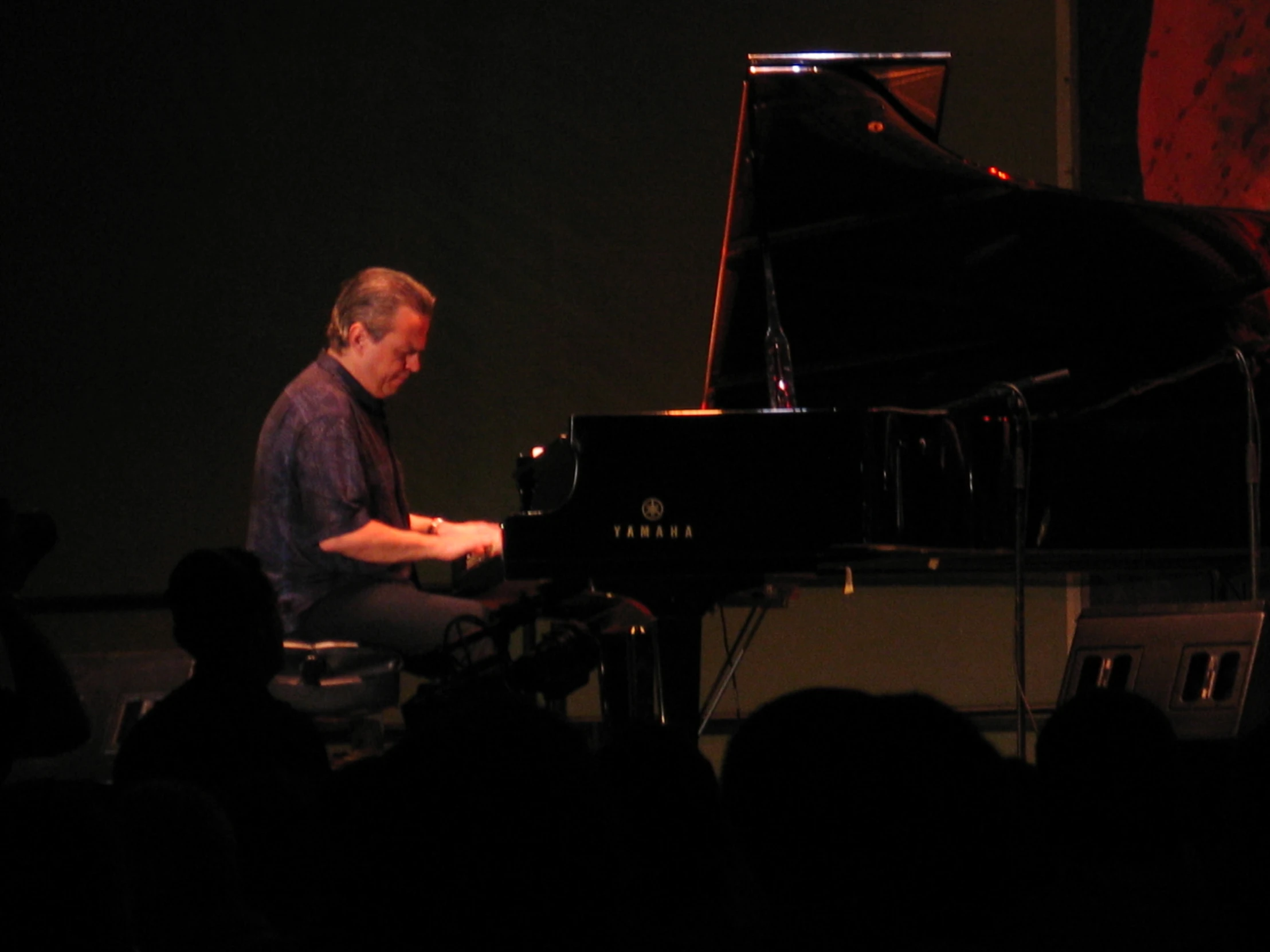 an older man sits at a black piano in front of a crowd