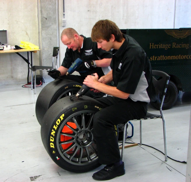 two men sitting on chairs while repairing the tires of some racing cars