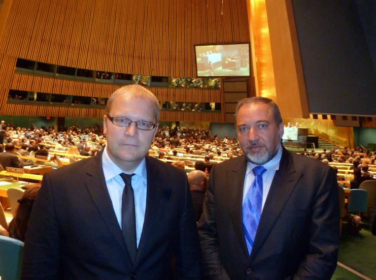 two men in suits stand next to each other in an auditorium