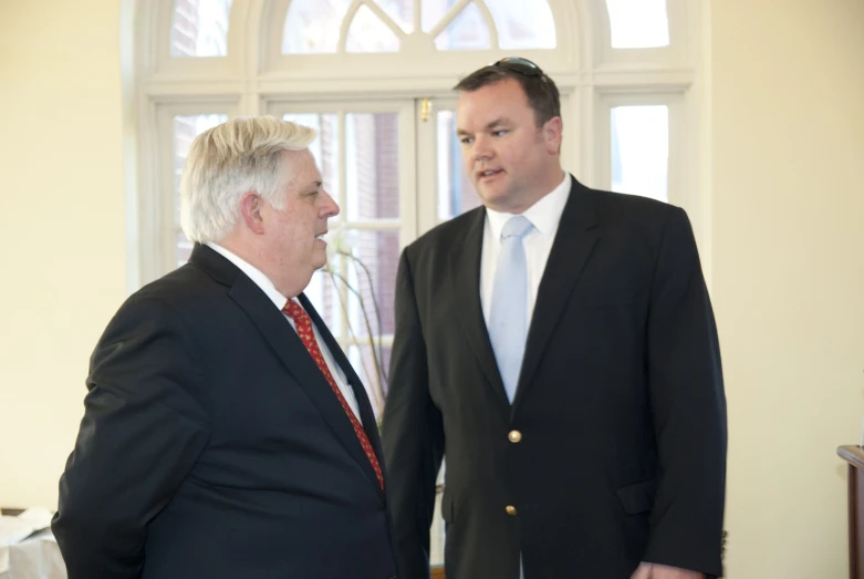 two men wearing suits standing side by side in front of windows