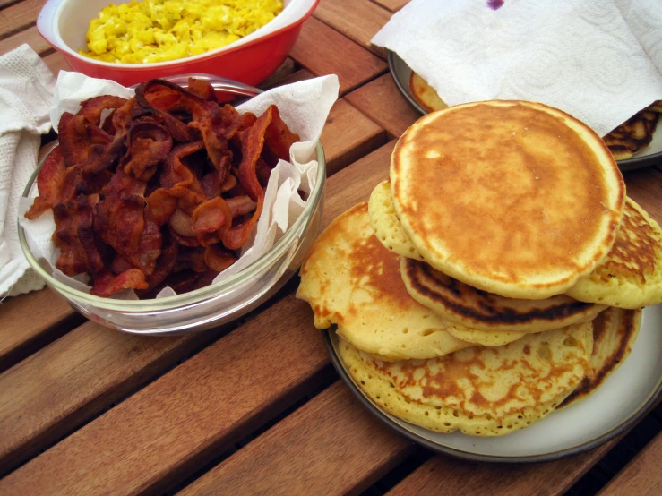 a wooden table holding two plates of pancakes and two bowls of scrambled eggs