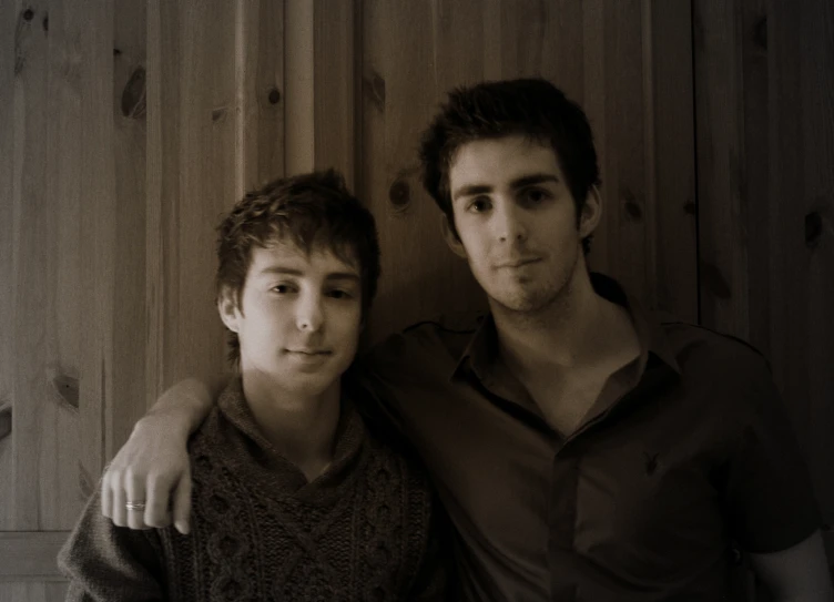 two men pose for the camera in front of wooden walls